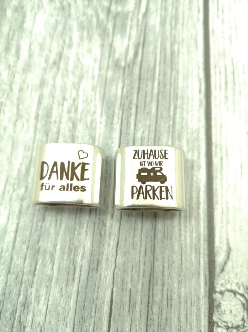 May include: Two silver metal charms with engraved text. The first charm says "Danke f&uuml;r alles" with a heart symbol. The second charm says "Zuhause ist wo wir parken" with a camper van symbol.