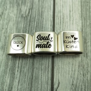 May include: Three silver metal charm bracelets with engraved text. The first charm says "Gl&uuml;ck" with a four-leaf clover symbol. The second charm says "Soulmate" with a small anchor symbol. The third charm says "Im K&uuml;sten Kind" with a heart symbol.