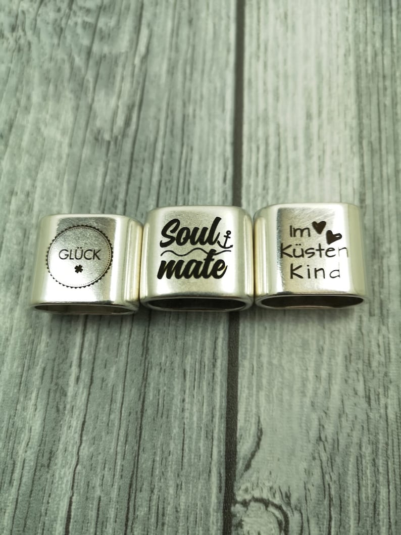 May include: Three silver metal charm bracelets with engraved text. The first charm says "Gl&uuml;ck" with a four-leaf clover symbol. The second charm says "Soulmate" with a small anchor symbol. The third charm says "Im K&uuml;sten Kind" with a heart symbol.