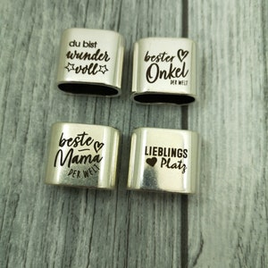 May include: Four silver metal charms with engraved text and hearts. The charms read "du bist wunder voll", "bester Onkel der Welt", "beste Mama der Welt", and "Lieblings Platz".