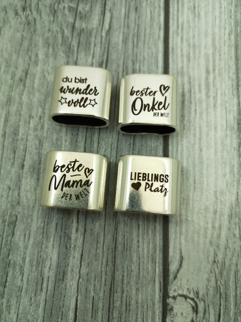 May include: Four silver metal charms with engraved text and hearts. The charms read "du bist wunder voll", "bester Onkel der Welt", "beste Mama der Welt", and "Lieblings Platz".