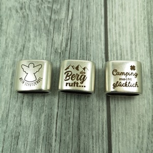 May include: Three silver metal keychains with engraved designs. The first keychain has an angel with the text "Mein Schutzengel". The second keychain has mountains with the text "Der Berg ruft...". The third keychain has a four-leaf clover with the text "Camping macht gl&uuml;cklich".