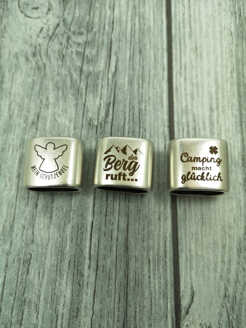 May include: Three silver metal keychains with engraved designs. The first keychain has an angel with the text "Mein Schutzengel". The second keychain has mountains with the text "Der Berg ruft...". The third keychain has a four-leaf clover with the text "Camping macht gl&uuml;cklich".