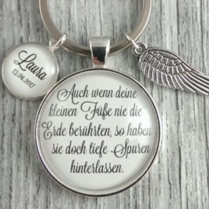 Könnte beinhalten: Ein silberner Schlüsselanhänger mit einem Glasanhänger, auf dem "Auch wenn deine kleinen Füße nie die Erde berührten, so haben sie doch tiefe Spuren hinterlassen." steht. Der Anhänger ist an einem silbernen Ring befestigt, an dem ein kleinerer silberner Anhänger mit der Aufschrift "Laura 13.04.2017" hängt. Ein silberner Engelsflügel-Anhänger ist ebenfalls am Ring befestigt.