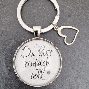 Pode incluir: Chaveiro de prata com um charme redondo de vidro que diz "Du bist einfach toll" em escrita preta. O charme está preso a um anel de prata com um charme de coração de prata.