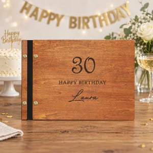 May include: A rectangular wooden photo album with a dark brown wood grain finish. The front cover features the number 30, the text HAPPY BIRTHDAY, and the name Laura engraved in black. A black vertical spine is secured with four gold-toned metal rivets.