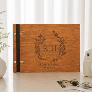 Könnte beinhalten: Ein rechteckiges Fotoalbum aus Holz mit dunkelbrauner Oberfläche und einem schwarzen vertikalen Buchrücken, der mit vier Metallnieten befestigt ist. Der Einband ist mit einem floralen Kranz, den Initialen R H, ROSE & HENRY und 16.10.2025 graviert.