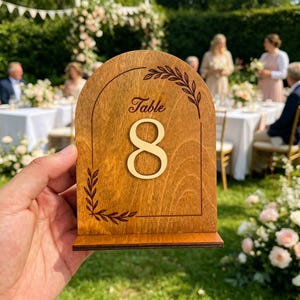 Könnte beinhalten: Holz-Tischkarte mit der goldenen Zahl acht. Das Schild hat einen dunkelbraunen Rahmen mit dem Wort "Table" und dekorativen Blättern. Das Schild wird bei einer Outdoor-Veranstaltung gezeigt.