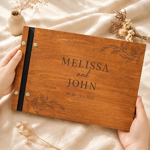 Könnte beinhalten: Ein rechteckiges Fotoalbum aus Holz mit dunkelbrauner Oberfläche und einem schwarzen Buchrücken, der mit vier silbernen Metallschrauben befestigt ist. Der Einband ist mit dem Text MELISSA and JOHN und 30. October 2025 sowie Blattmotiven graviert.