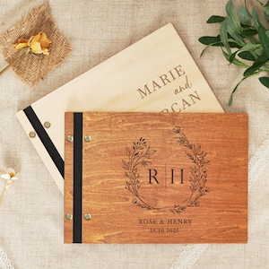 Könnte beinhalten: Zwei Gästebücher aus Holz. Eines ist hell mit den eingravierten Namen "Marie und CAN". Das andere ist dunkleres Holz mit floralem Kranzdesign, den Initialen "R H" und den Namen "Rose & Henry" und dem Datum "16.10.2025".