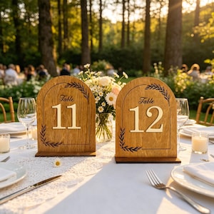 Könnte beinhalten: Zwei Tischschilder aus Holz mit den Nummern "Tisch 11" und "Tisch 12" werden auf einer weißen Tischdecke angezeigt. Die Schilder haben eine gewölbte Oberseite mit dekorativen Blättern. Eine Vase mit Blumen und Weingläsern steht ebenfalls auf dem Tisch.