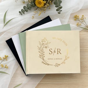 Könnte beinhalten: Eine Auswahl an Hochzeitsgästebüchern in verschiedenen Farben, darunter Schwarz, Weiß und Salbeigrün. Das vordere Buch ist cremefarben mit goldfarbenem Text "S & R" in einem floralen Kranz, dem Datum "08.July.2026" und den Namen "Sophia & Ronald".