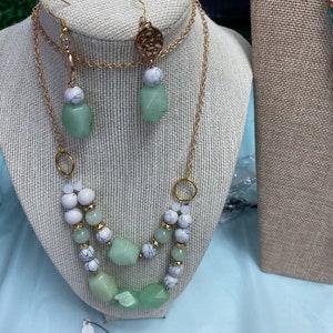 May include: A gold chain necklace with green and white beads. The necklace is layered and has a pendant with a gold design. Matching green and white earrings are also included.