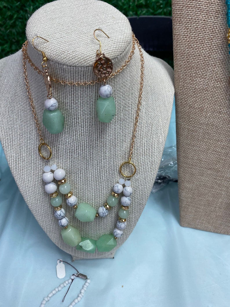 May include: A gold chain necklace with green and white beads. The necklace is layered and has a pendant with a gold design. Matching green and white earrings are also included.