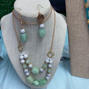 May include: A two-strand necklace with green and white beads and a matching pair of earrings. The necklace has a gold chain and the earrings have a gold charm.