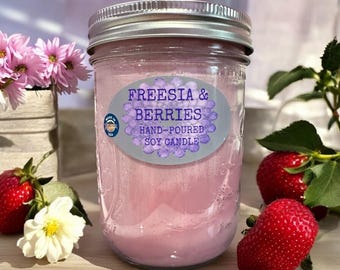 May include: A pink soy candle in a clear glass jar with a silver lid. The label reads "Freesia & Berries Hand-Poured Soy Candle". The candle is surrounded by strawberries and flowers.