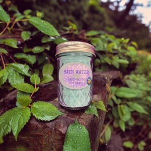 May include: A teal green soy candle in a glass jar with a pink lid. The label reads "Rain Water Hand-Poured Soy Candle".