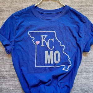 Puede incluir: Una camiseta azul real con un contorno plateado brillante del estado de Misuri. Dentro del contorno está el texto "KC MO" con un corazón rosa en el centro.
