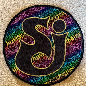 May include: Circular patch with a black background and a gold outline of the letters "SJ". The border is a rainbow pattern with a reptile skin texture. The patch is surrounded by a black border.
