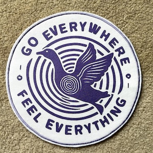May include: Circular patch with a white background and a purple duck illustration. The duck is flying within concentric circles. The text "GO EVERYWHERE - FEEL EVERYTHING" is printed around the edge.