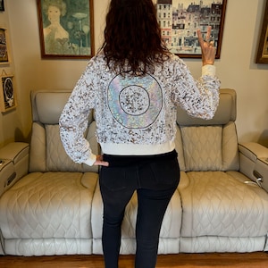 May include: A white sequined jacket with a large, iridescent circle design on the back. The jacket has a white collar and cuffs. The person in the image is wearing black jeans. The background includes framed artwork and a light-colored sofa.