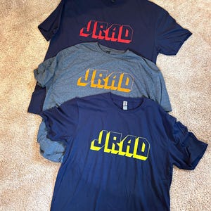 May include: Three t-shirts with the word "JRAD" printed on them in a 3D block style. The top shirt is navy blue with red lettering, the middle shirt is gray with orange lettering, and the bottom shirt is navy blue with yellow lettering.