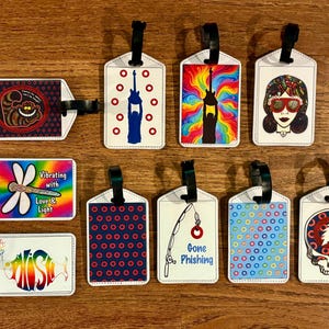May include: A collection of ten rectangular luggage tags with black straps, featuring various graphic designs including a cat, guitars, a person with sunglasses, a dragonfly with the text Vibrating with Love & Light, a fish, donuts, and a skull.