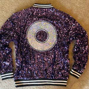 May include: A purple sequined jacket with a large silver circle design on the back. The jacket has black and white striped trim on the collar, cuffs, and hem. The jacket is a bomber style.