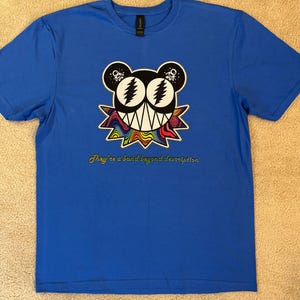 May include: A bright blue short-sleeve t-shirt featuring a central graphic of a stylized bear head with lightning bolt eyes, a jagged mouth, and a rainbow-colored collar. Below the graphic, the text reads, They're a band beyond description.