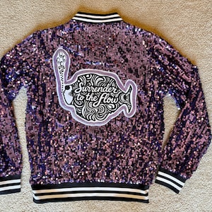 May include: A purple sequined jacket with a black and white striped collar, cuffs, and hem. The back of the jacket features a white and black graphic with the words "Surrender to the Flow".