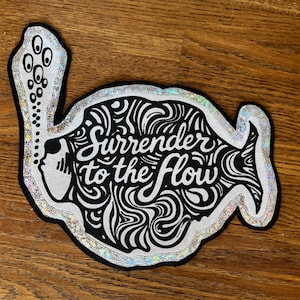 May include: A black and white patch in the shape of a fish with the words "Surrender to the Flow". The patch has a silver glitter border and is on a wooden surface. The design features a woman's profile and swirling patterns.