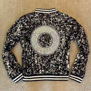 May include: Black sequin bomber jacket with a large, iridescent circle design on the back. The jacket has a white and black striped collar, cuffs, and hem. The sequins create a shimmering effect.