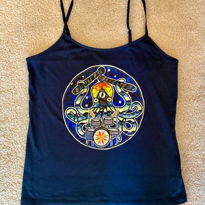 May include: A dark blue tank top featuring a circular graphic print of an octopus playing a drum set. The octopus is rendered in a stained-glass style with vibrant blue, green, and yellow colors, holding drumsticks above a multi-piece drum kit.