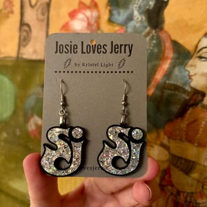 May include: A pair of dangle earrings with a black outline and filled with silver glitter. The earrings are in the shape of the letters "J" and "J" with a decorative swirl. The earrings are displayed on a gray card that reads "Josie Loves Jerry" and "by Kristel Light".