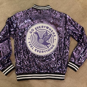 May include: A purple sequined bomber jacket with black and white striped trim. The back of the jacket features a circular patch with a purple bird graphic and the words "GO EVERYWHERE FEEL EVERYTHING".