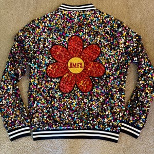 May include: A multi-colored sequined bomber jacket with a large red flower design on the back. The flower has a yellow center with the letters "BMF5". The jacket has black and white striped trim on the collar, cuffs, and hem.