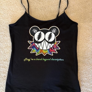 May include: A black spaghetti strap tank top featuring a graphic of a stylized bear head with lightning bolt eyes and a rainbow-colored collar. Below the graphic, the text reads, "They're a band beyond description" in a cursive, multi-colored font.