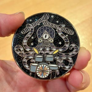 May include: A round, black enamel pin with a silver outline. The pin features a purple octopus with a third eye playing a drum set against a black background with silver glitter and white stars.