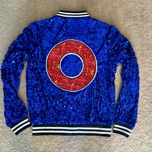 May include: A blue sequined jacket with a large red and white circular design on the back. The jacket has a black and white striped collar, cuffs, and hem. The jacket is a vibrant, eye-catching piece.