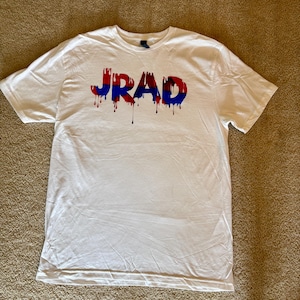 May include: White t-shirt with the word "JRAD" in red and blue dripping letters. The shirt is made of a soft material and has a crew neck. The shirt is laying on a brown surface.