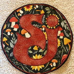 May include: A round, decorative fabric item featuring a large, red letter "S" with a dotted pattern. The background is black with a pattern of mushrooms, sunflowers, and leaves. The item has a black border.