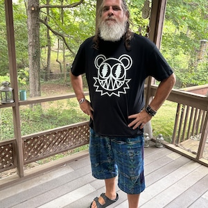 May include: A man wearing a black t-shirt with a white graphic of a bear with lightning bolt eyes. He is wearing blue patterned shorts and black sandals. He is standing on a wooden porch.