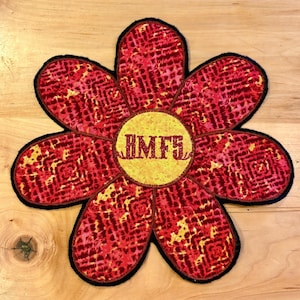 May include: A flower-shaped coaster with a red and yellow tie-dye pattern. The coaster has a black border and a yellow center with the letters "HMFS" in red. The coaster is likely made of fabric.
