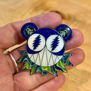 May include: Enamel pin featuring a stylized bear design with a blue head, white eyes, and a jagged, colorful lower section. The bear's ears have astronaut details. The pin has a silver border and a secure pin back.