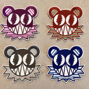 May include: Four embroidered bear patches in various colors: purple, red, brown, and blue. Each bear has white teeth, eyes, and a jagged, starburst-like design around the face. The bear ears have striped details.