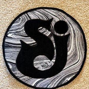 May include: A round patch featuring a black "S" and "J" design. The background has a marbled effect in shades of gray and white. The patch has a black border.