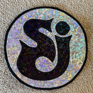 May include: Circular patch with a black border. The center features the letters "SJ" in a stylized black font, set against a shimmering, iridescent background. The patch has a holographic, textured appearance.