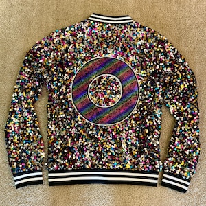 May include: A black sequined bomber jacket with a large, rainbow-colored circular design on the back. The jacket is covered in colorful sequins and features black and white striped trim on the collar, cuffs, and hem.