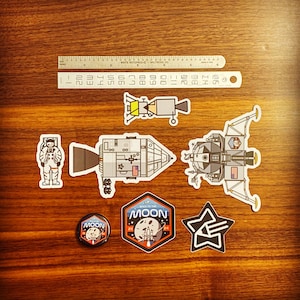 Könnte beinhalten: Ein Set mit Weltraum-Stickern und einem Knopf. Die Sticker zeigen ein Mondmodul, einen Astronauten, eine Rakete, einen Stern und einen Patch mit der Aufschrift "Zurück zum Mond 2024". Der Knopf ist rund und zeigt einen Mond mit dem Wort "Mond" darauf.
