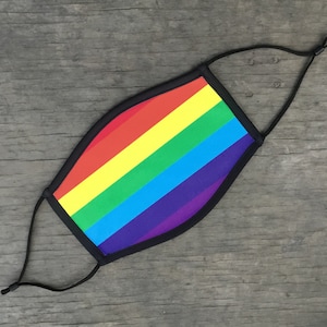 May include: A rainbow-colored face mask with black elastic ear loops. The mask is made of a soft, breathable fabric and features a rainbow design with red, orange, yellow, green, blue, indigo, and violet stripes.